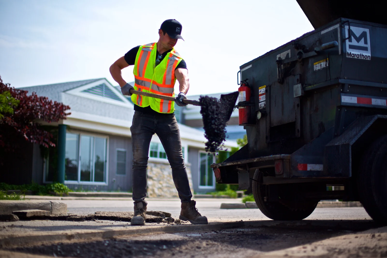 A crew member shovels hot asphalt beside a truck during an asphalt patching job